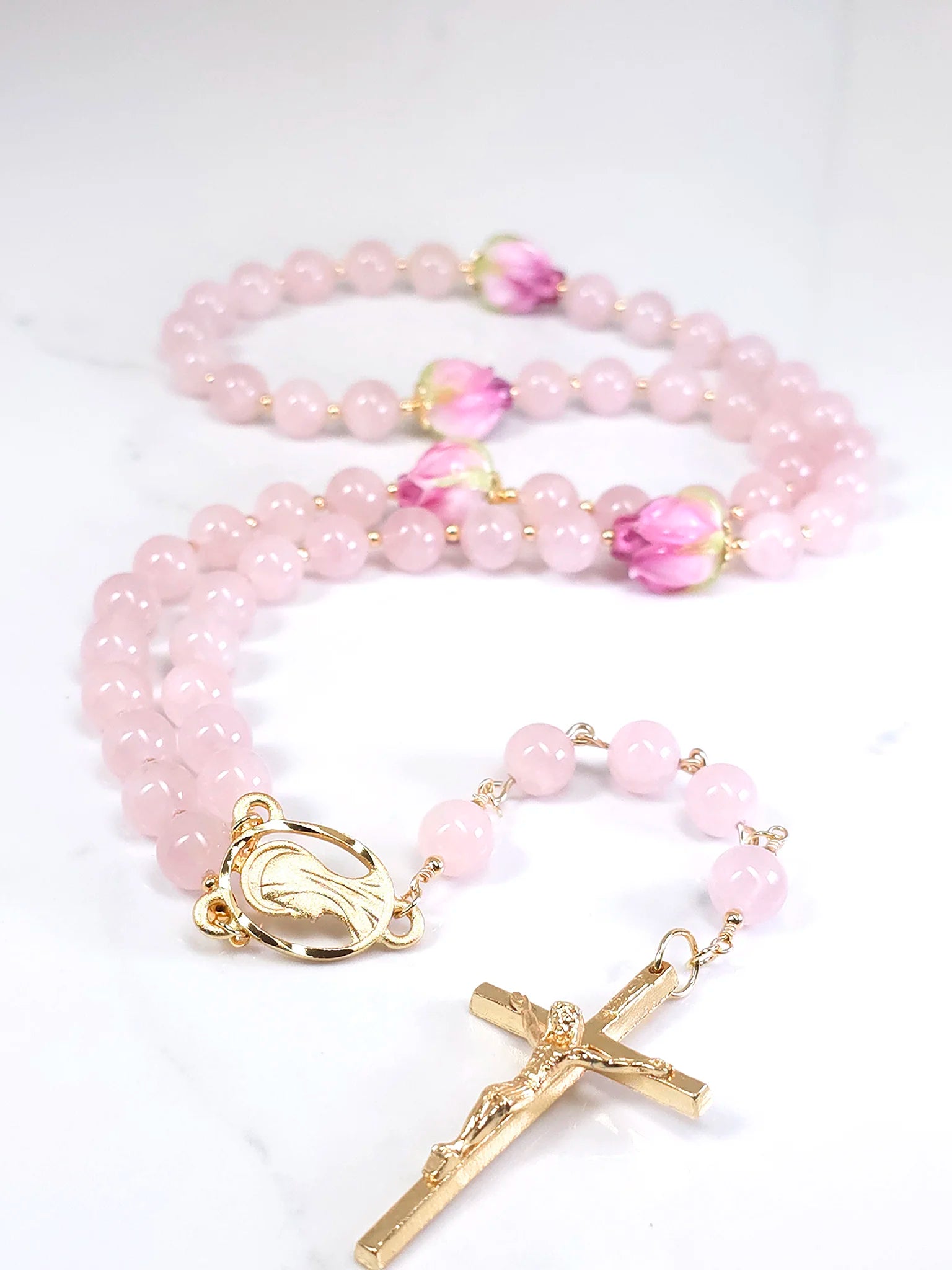 Delicate necklace rosary crafted from pink quartz with a gold-plated Crucifix pendant, resting on a white table.