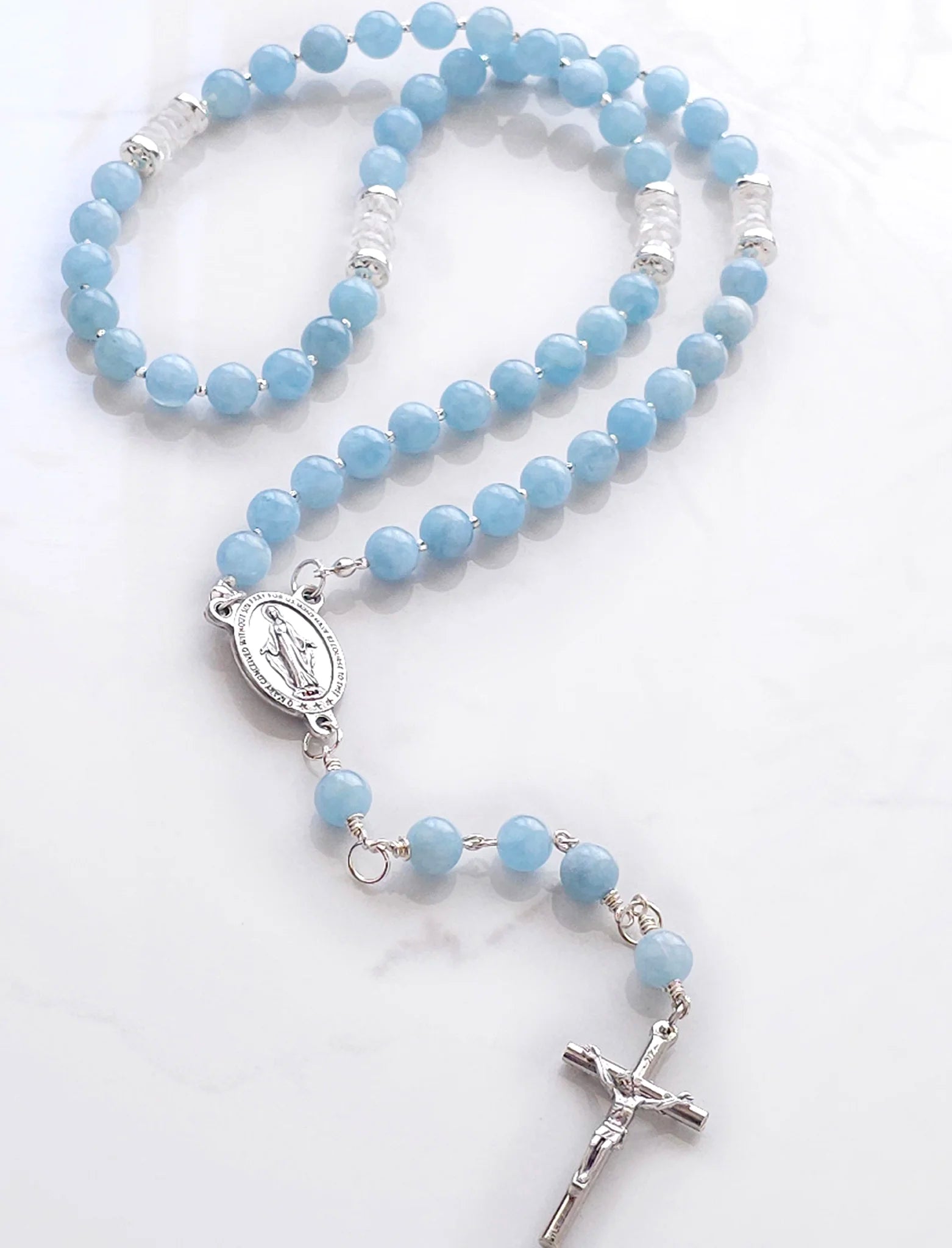 Blue rosary with natural Aquamarine beads, complemented by Sterling Silver spacers, laid on a white table.