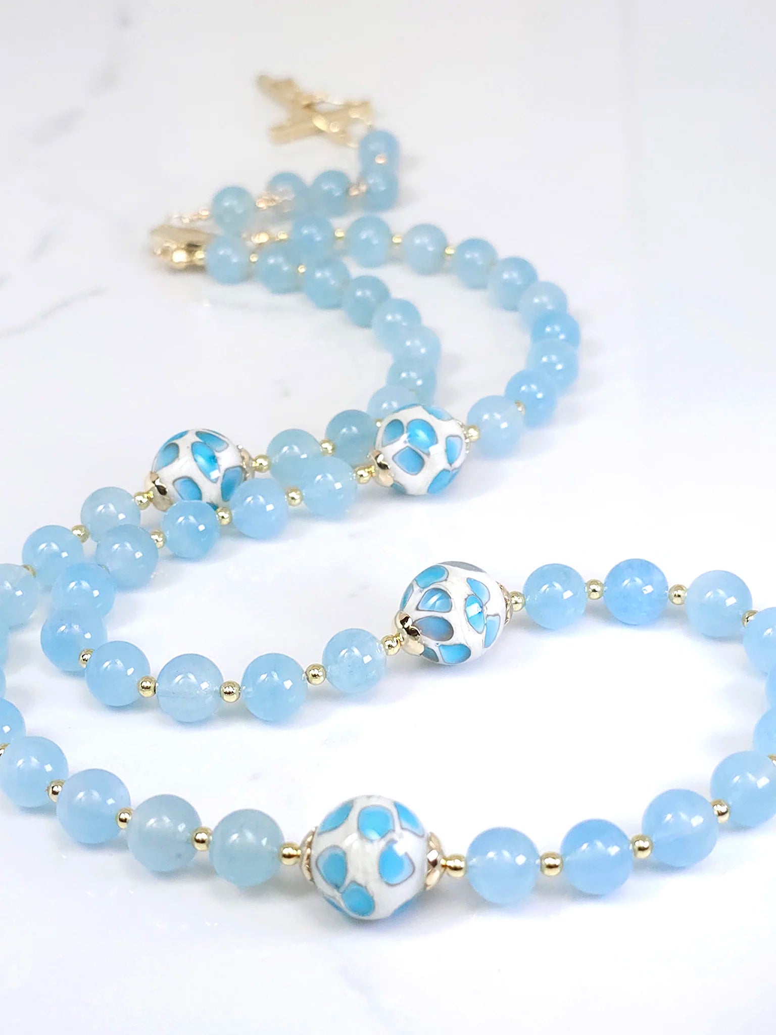 Rosary necklace adorned with intricate beads and blue little flower glass bead accents, laid out on a white table backdrop.