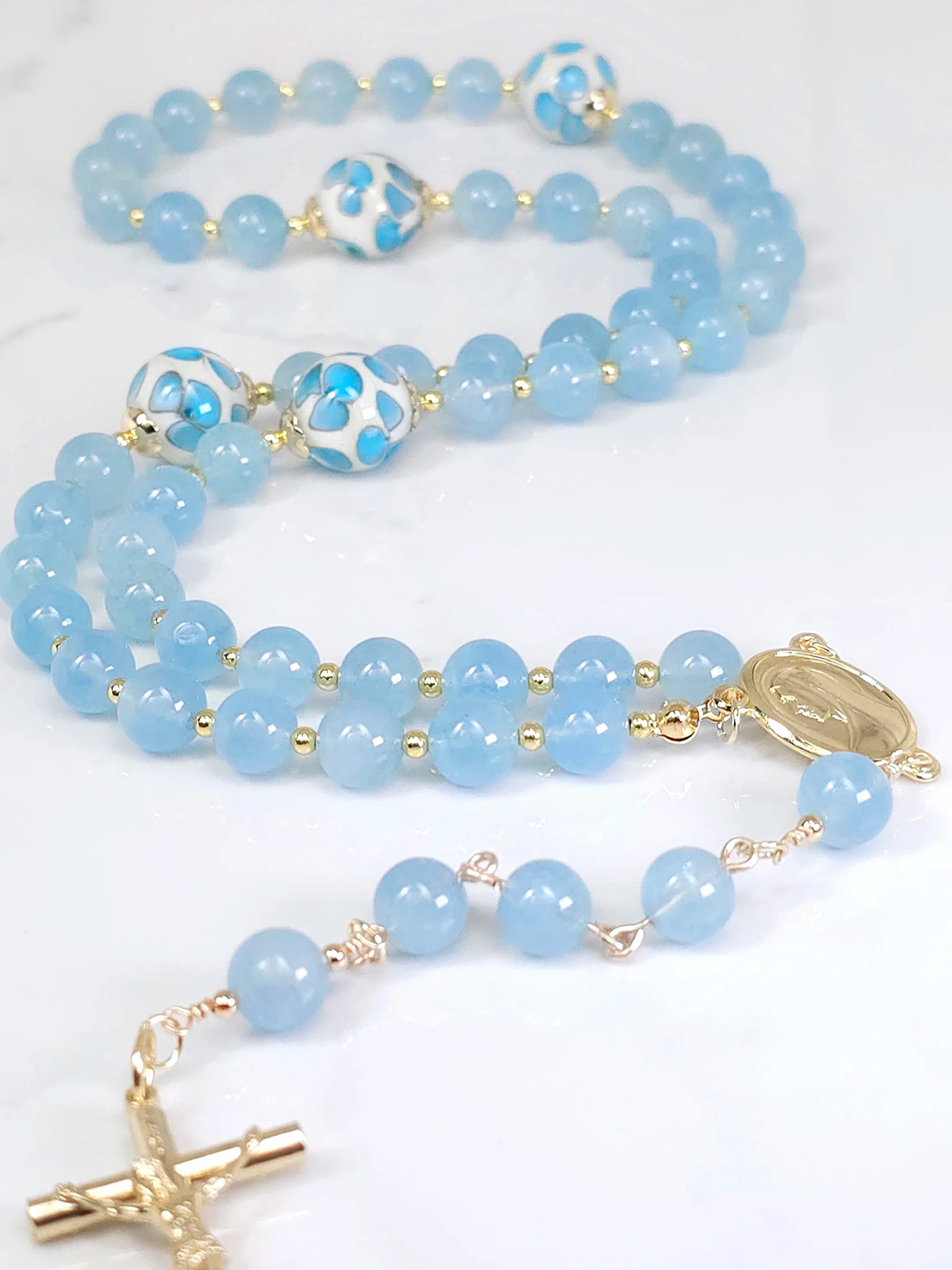 Necklace rosary made of shimmering beads, highlighted by a Gold-plated Madonna medal centerpiece, draped on a white table.