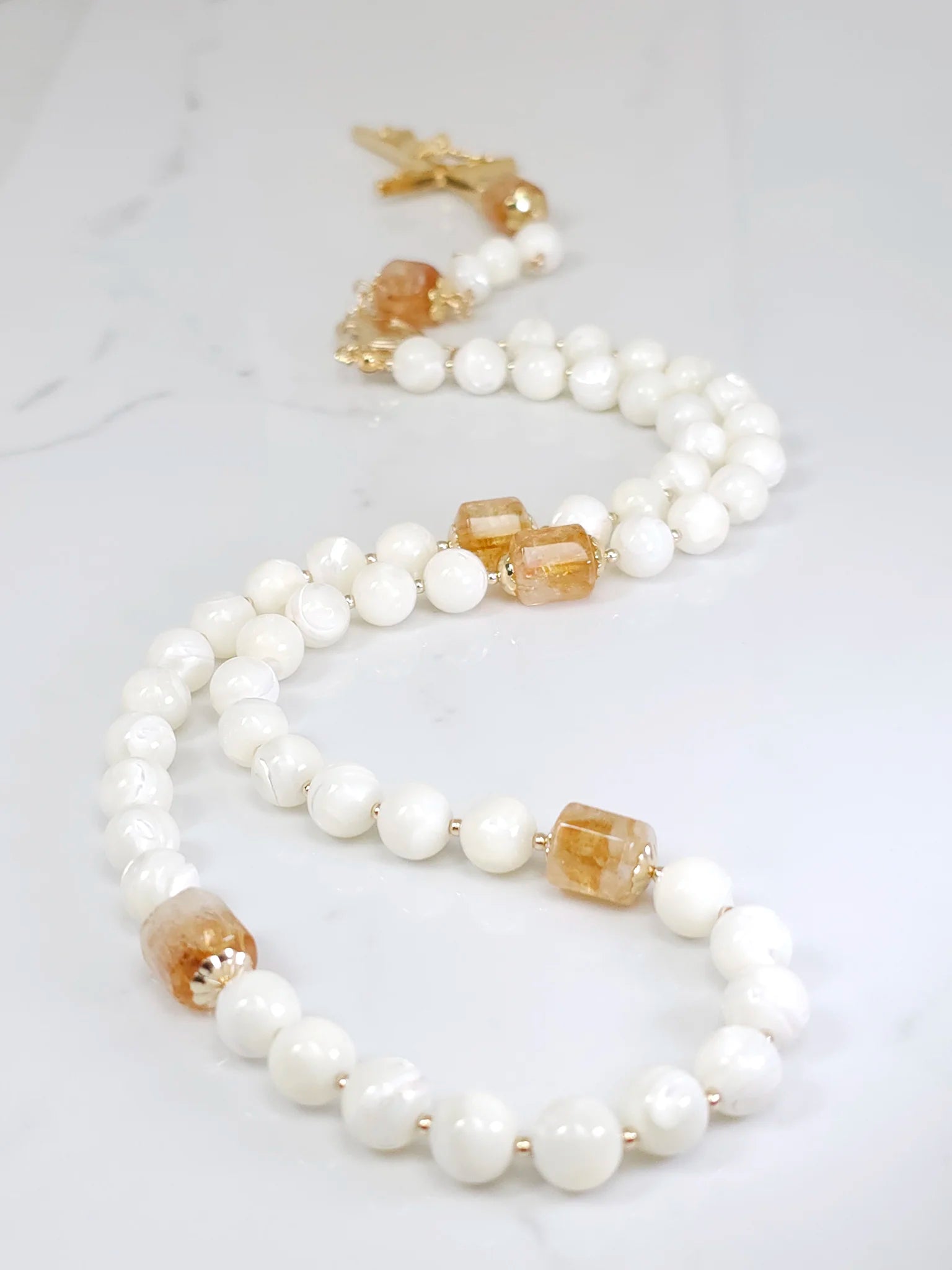Rosary necklace blending of Mother of Pearl with luminous Citrine, featuring a Crucifix pendant, on a white table.