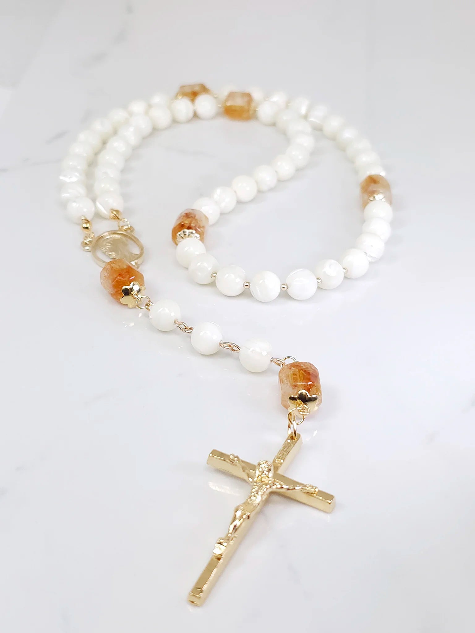Rosary necklace combining White Mother of Pearl and sunlit Citrine, adorned with a Madonna medal, laid on a white table.