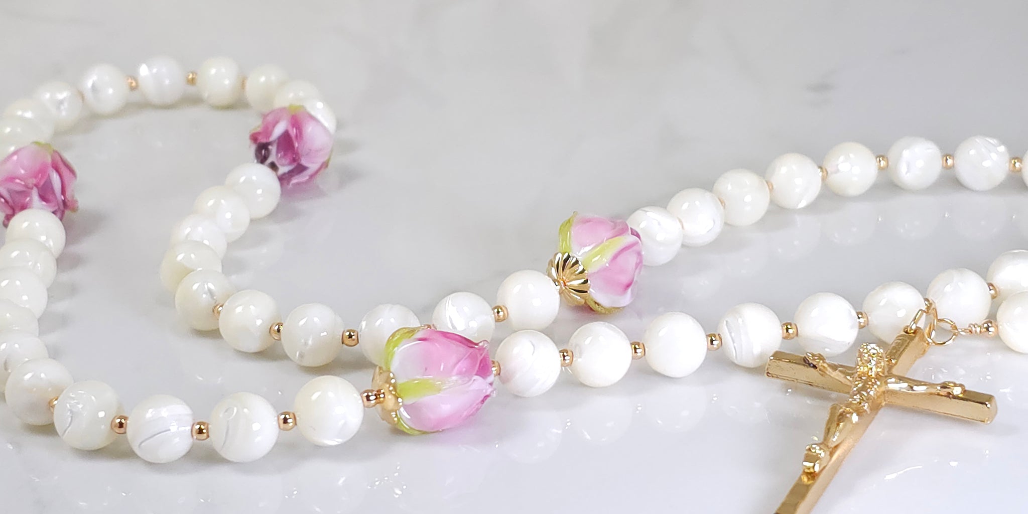 White Mother of Pearl Catholic Rosary Beads with Gold Cross and Glass Rose buds beads placed on top of white marble.
