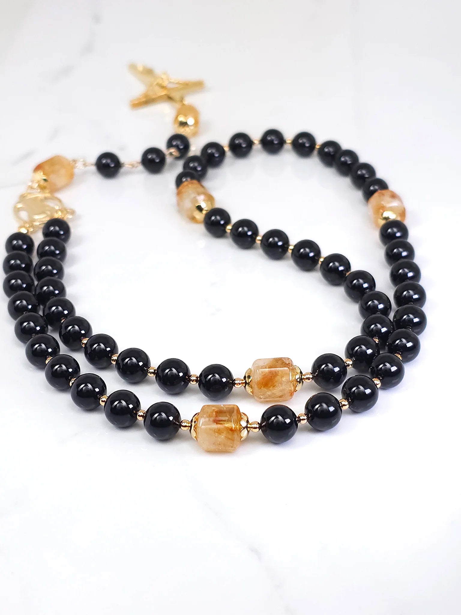 Elegant rosary combining Black Onyx beads with Golden Citrine, complemented by a Gold Madonna medal, laid on a white table.