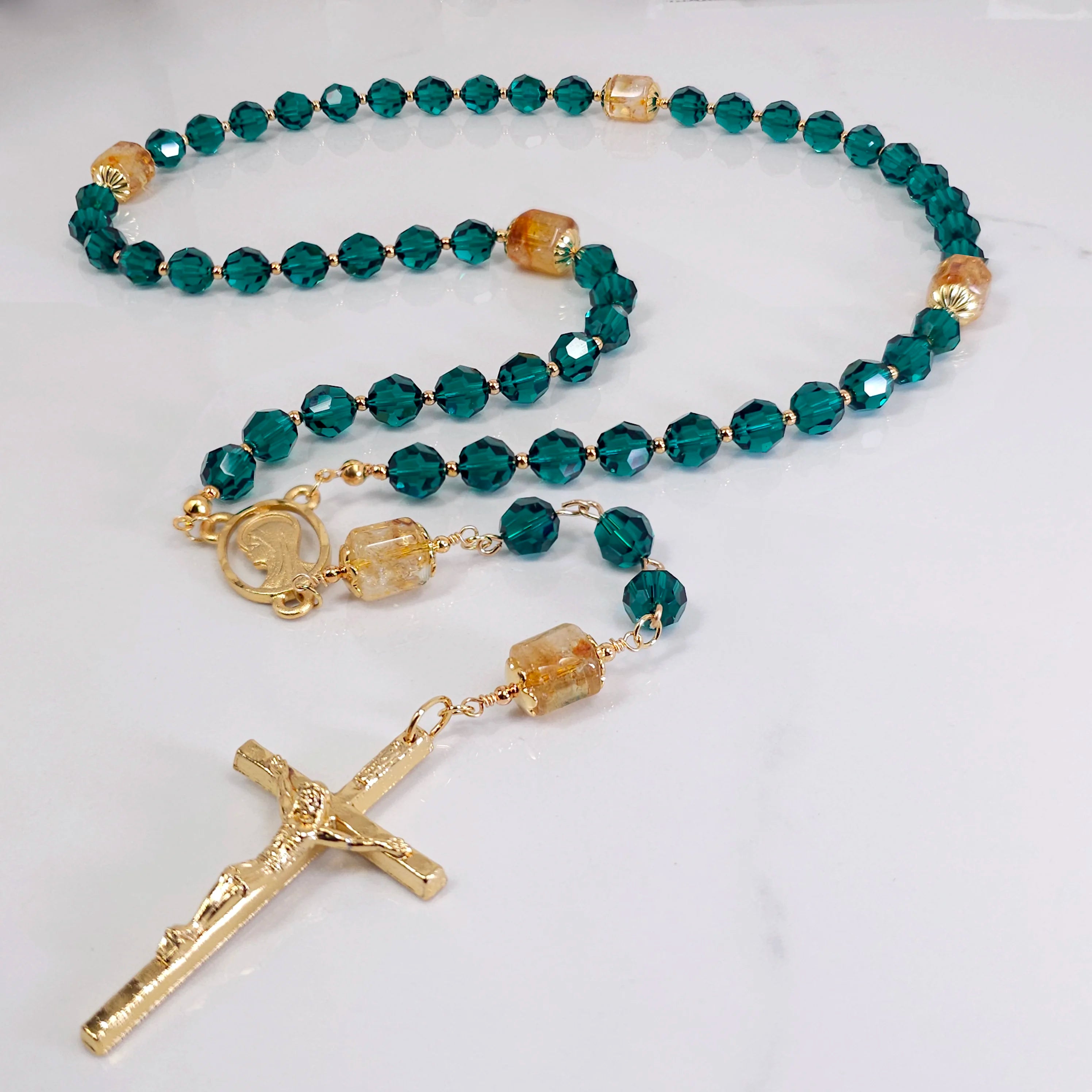 Green rosary made from Swarovski crystal embellished with gold cross and Madonna medal, lying on white marble.